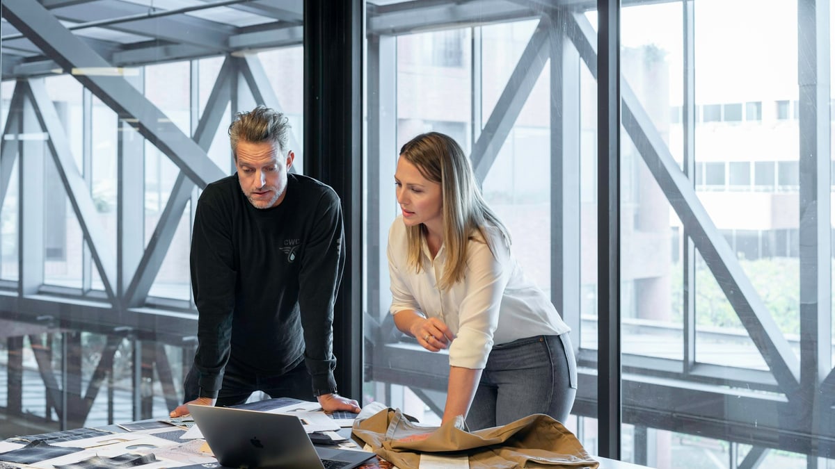 Two colleagues leaning over a table of printed photographs and a laptop, reviewing creative layouts