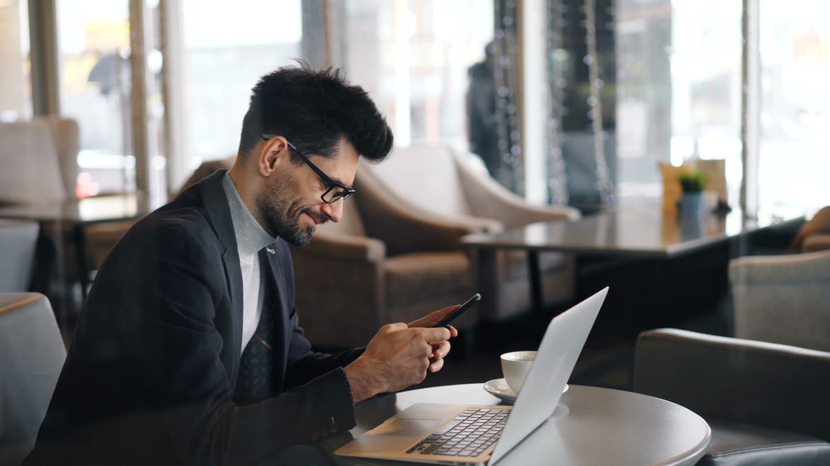 A man in a suit using a smartphone beside an open laptop in a cafe