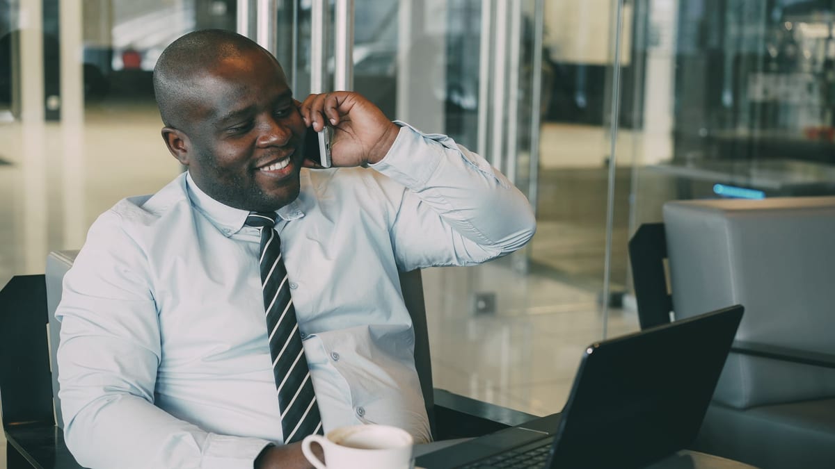 A man on a phone call at an open laptop in a glass-walled office