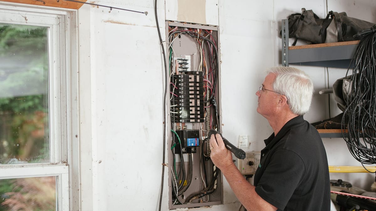 An electrician in a black shirt checking a home fuse box with a tester